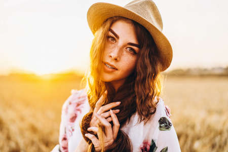 Portrait of a beautiful young woman with curly hair and freckles face. Woman in dress and hat posing in wheat field at sunset and looking at camera.の写真素材