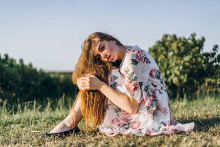 Full length portrait of beautiful woman with long curly hair on currant field background. Girl in a light dress sits on the grass in sunny day.の写真素材
