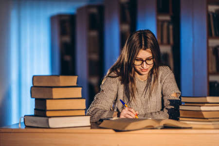 Young student in glasses preparing for the exam. Girl in the evening sits at a table in the library with a pile of books.の写真素材