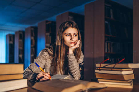 Young student in glasses preparing for the exam. Girl in the evening sits at a table in the library with a pile of books.の写真素材