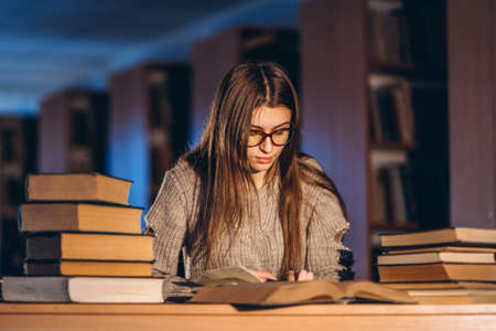 Young student in glasses preparing for the exam. Girl in the evening sits at a table in the library with a pile of books.の写真素材