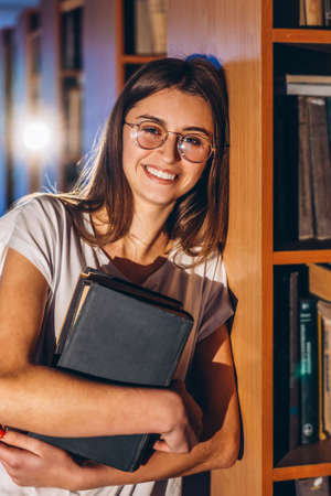 Young girl student with glasses in library stands near bookshelves. Girl smiles and holds a book in her hands. Exam preparation.の写真素材