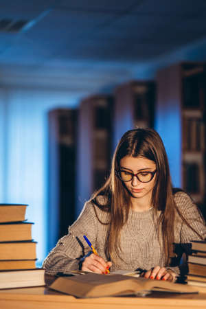 Young student in glasses preparing for the exam. Girl in the evening sits at a table in the library with a pile of books.の写真素材