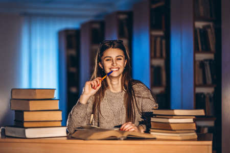 Young happy smiling student in glasses preparing for the exam. Girl in the evening sits at a table in the library with a pile of books, smiling and looking at the camera.の写真素材