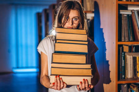 Young girl student with glasses in library smiling and carries stack of books. Exam preparation.の写真素材