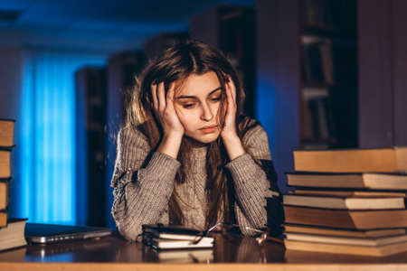 Tired student girl in the evening sitting leaning on his arm in the library at the desk with books. Teaching and preparation for exams.の写真素材