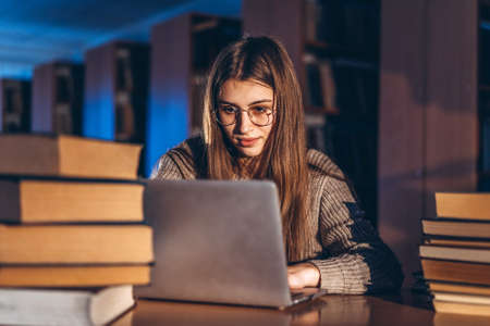 Young student woman in the evening sits at a table in the library with a pile of books and works on a laptop. Preparing for the exam.の写真素材