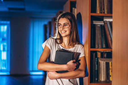 Young girl student with glasses in library stands near bookshelves. Girl smiles and holds a book in her hands. Exam preparation.の写真素材