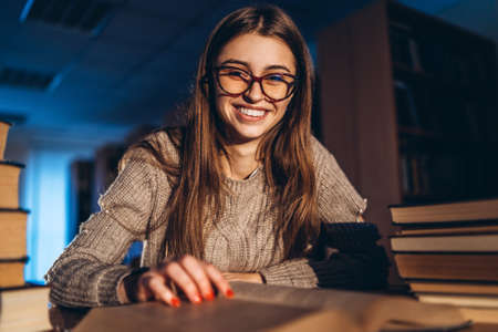 Young happy smiling student in glasses preparing for the exam. Girl in the evening sits at a table in the library with a pile of books, smiling and looking at the camera.の写真素材