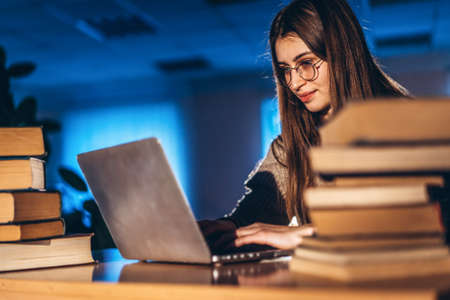 Young student woman in the evening sits at a table in the library with a pile of books and works on a laptop. Preparing for the exam.の写真素材