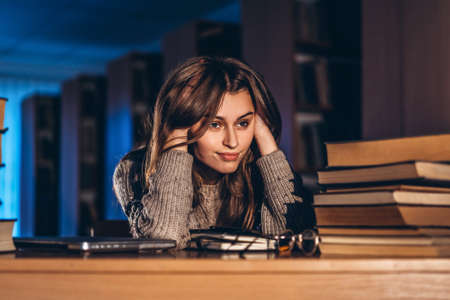 Tired student girl in the evening sitting leaning on his arm in the library at the desk with books. Teaching and preparation for exams.の写真素材