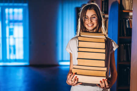 Young girl student with glasses in library smiling and carries stack of books. Exam preparation.の写真素材