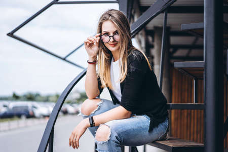 Beautiful girl with long hair and glasses sitting on metal stairs on the wooden background of house with vertical boards. Woman smiling and looking at camera.の写真素材
