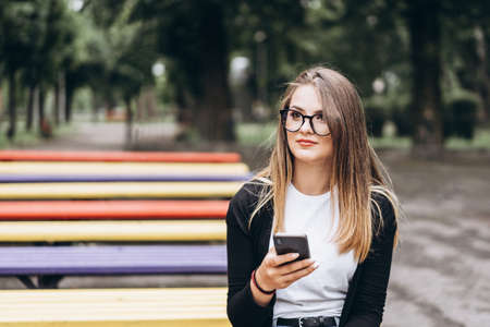 Young girl in glasses looks into a cellphone. Sitting on a park bench. Concept of communication and social networks.の写真素材