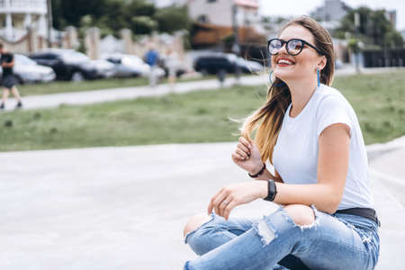 Young woman with long hair in stylish glasses sitting on the ground in the city park. Girl dressed in jeans and t-shirt smiling and looking away.の写真素材
