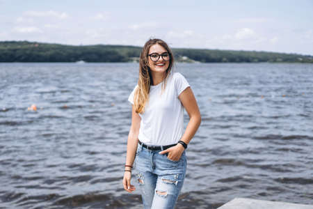 Portrait of a beautiful young woman in stylish glasses. Girl in white tshirt posing on the background of the lake landscape.の写真素材