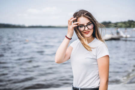 Portrait of a beautiful young woman in stylish glasses. Girl in white tshirt posing on the background of the lake landscape.の写真素材