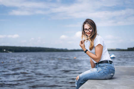 Young woman with long hair in stylish glasses posing on the concrete shore near the lake. Girl dressed in jeans and t-shirt smiling and looking away.の写真素材