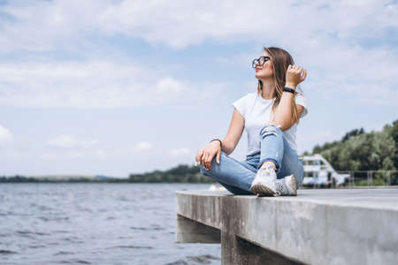 Young woman with long hair in stylish glasses posing on the concrete shore near the lake. Girl dressed in jeans and t-shirt smiling and looking away.の写真素材