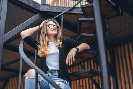 Beautiful girl with long hair and glasses sitting on metal stairs on the wooden background of house with vertical boards. Woman smiling and looking at camera.の写真素材