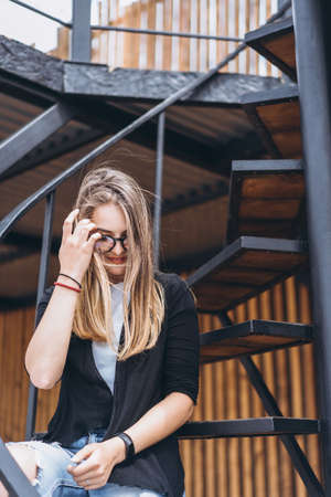 Beautiful girl with long hair and glasses sitting on metal stairs on the wooden background of house with vertical boards. Woman smiling and looking at camera.の写真素材