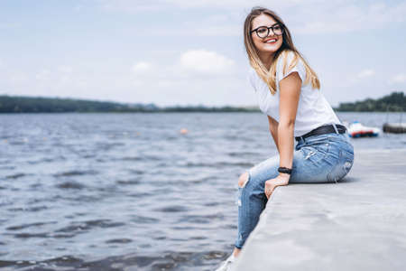 Young woman with long hair in stylish glasses posing on the concrete shore near the lake. Girl dressed in jeans and t-shirt smiling and looking away.の写真素材