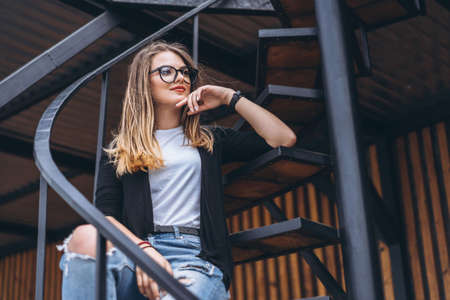 Beautiful girl with long hair and glasses sitting on metal stairs on the wooden background of house with vertical boards. Woman smiling and looking at camera.の写真素材