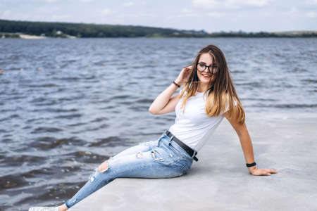 Young woman with long hair in stylish glasses posing on the concrete shore near the lake. Girl dressed in jeans and t-shirt smiling and looking at the camera.の写真素材