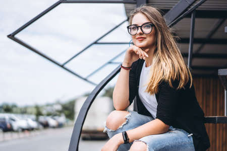 Beautiful girl with long hair and glasses sitting on metal stairs on the wooden background of house with vertical boards. Woman smiling and looking at camera.の写真素材