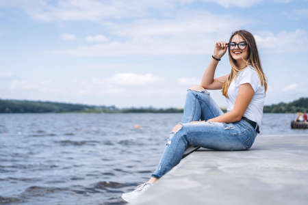 Young woman with long hair in stylish glasses posing on the concrete shore near the lake. Girl dressed in jeans and t-shirt smiling and looking at the camera.の写真素材