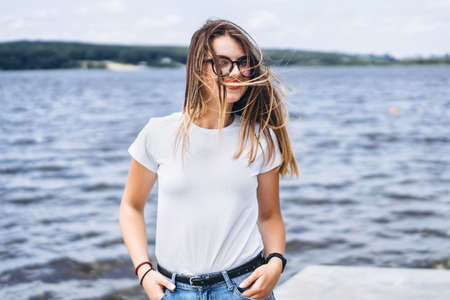 Portrait of a beautiful young woman in stylish glasses. Girl in white tshirt posing on the background of the lake landscape.の写真素材
