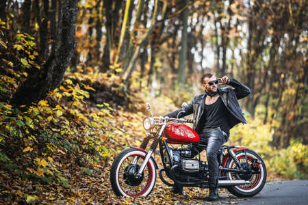 Bearded brutal man in sunglasses and leather jacket sitting on a motorcycle on the road in the forest with blured colorful background.の写真素材