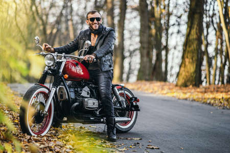 Bearded brutal man in sunglasses and leather jacket sitting on a motorcycle on the road in the forest with blured colorful background.の写真素材
