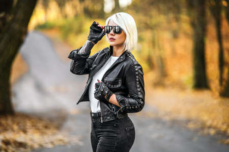 Pretty blonde biker styled women in black leather jacket with sunglasses standing outdoors in the forest with colorful blurred backgroundの写真素材