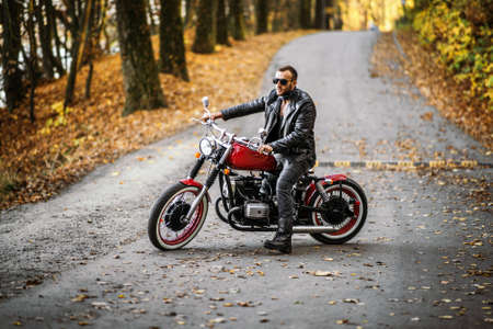 Bearded brutal man in sunglasses and leather jacket sitting on a motorcycle on the road in the forest with blured colorful background.の写真素材