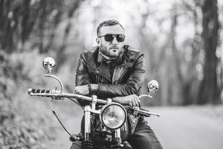 Bearded brutal man in sunglasses and leather jacket sitting on a motorcycle on the road in the forest with blured colorful background.の写真素材