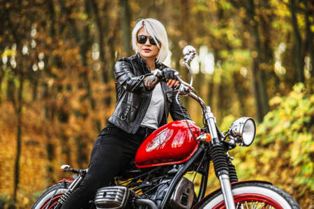 Pretty blonde biker girl in sunglasses with red motorcycle on the road in the forest with colorful blurred background.の写真素材