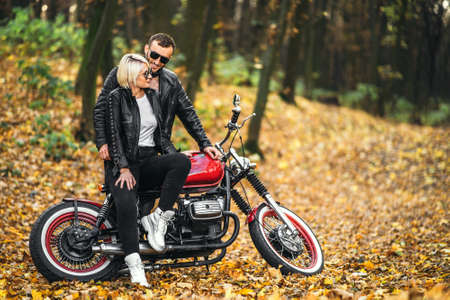 Pretty couple near red motorcycle on the road in the forest with colorful blured background. Relationship concept.の写真素材
