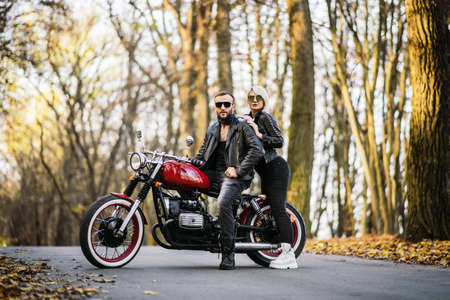 Pretty couple near red motorcycle on the road in the forest with colorful blured background. Relationship concept.の写真素材