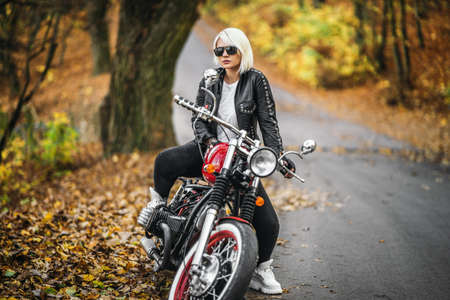 Pretty blonde biker girl in sunglasses with red motorcycle on the road in the forest with colorful blurred background.の写真素材