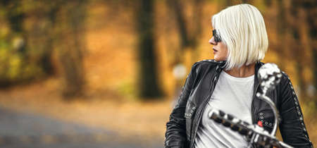 Pretty blonde biker girl in sunglasses with red motorcycle on the road in the forest with colorful blurred background.の写真素材