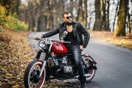 Bearded brutal man in sunglasses and leather jacket sitting on a motorcycle on the road in the forest with blured colorful background.の写真素材
