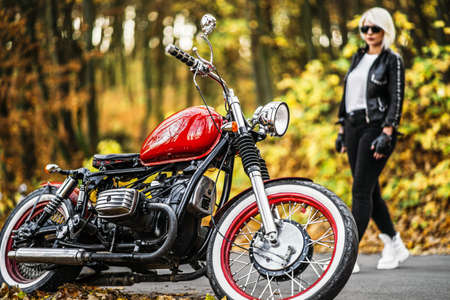 Pretty blonde biker girl in sunglasses with red motorcycle on the road in the forest with colorful blurred background.の写真素材