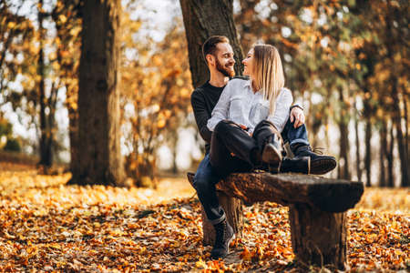 A young loving couple sitting on a wooden bench in the forest. Man and woman hugging and smiling on the background of autumn trees.の写真素材