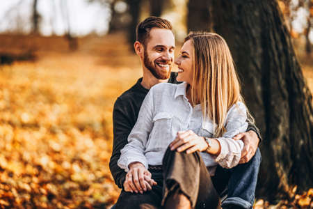 A young loving couple sitting on a wooden bench in the forest. Man and woman hugging and smiling on the background of autumn trees.の写真素材