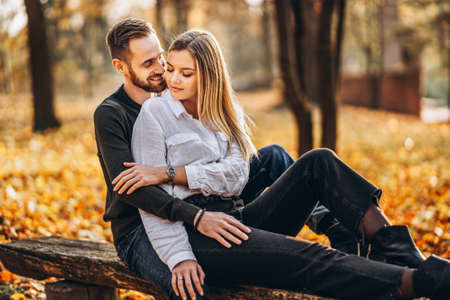 A young loving couple sitting on a wooden bench in the forest. Man and woman hugging and smiling on the background of autumn trees.の写真素材