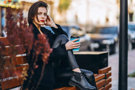 Young woman with short haircut, dressed in black, sits on a bench in the city. A woman holds a cup of coffee and looks into the camera.の写真素材