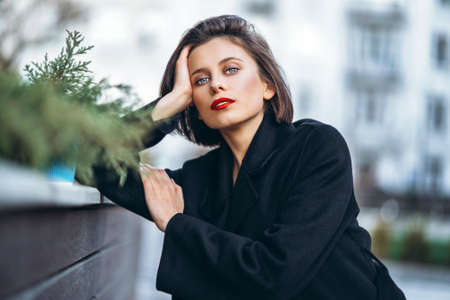 Close up portrait of young woman with short haircut and red lips, dressed in black. Woman posing, leaning on a wooden fence with plants. Blurred urban background.の写真素材