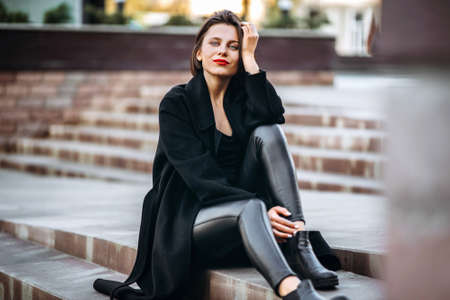 Full length portrait of young woman with short haircut and red lips, dressed in black. Woman sitting on the stairs outdoors on the stairsの写真素材