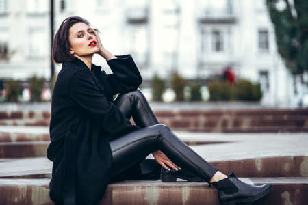 Full length portrait of young woman with short haircut and red lips, dressed in black. Woman sitting on the stairs outdoors on the stairsの写真素材